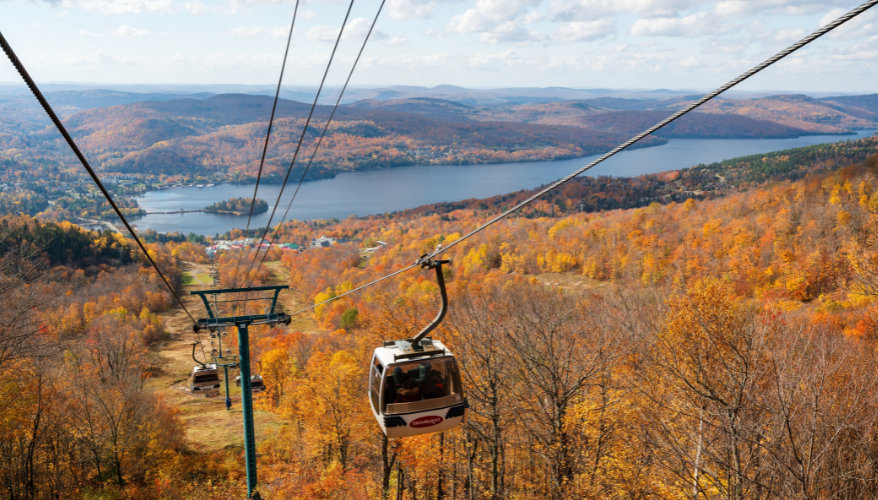 The summit of Mont-Tremblant Mont-Tremblant gondola flying over an autumn-coloured forest with views of the mountains and Lake Tremblant.