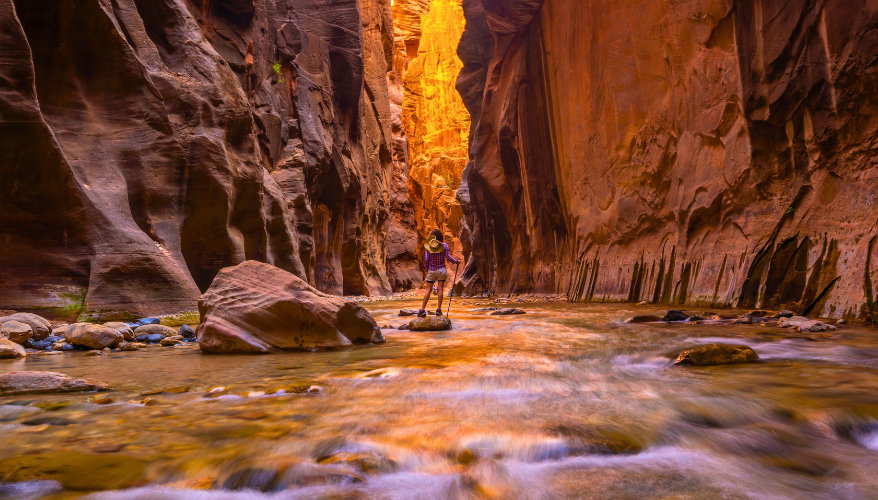 The Narrows A view of the trail with a woman standing with her back to the camera on a rock, surrounded by river water and high rock walls.