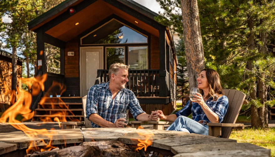 A couple relaxes around a campfire in front of their private cabin at Explorer Cabins at Yellowstone, at the gates of Yellowstone National Park. © Photo Explorer Cabins at Yellowstone Couple enjoying a campfire in front of a wooden cabin at Explorer Cabins at Yellowstone